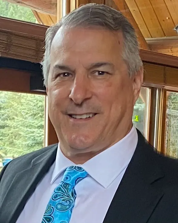 Brett Hightower: Smiling man with gray hair in a dark jacket and blue paisley tie, seated near a window