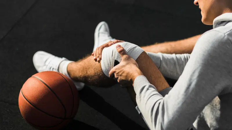A young man sits on a court beside a basketball, holding his injured knee