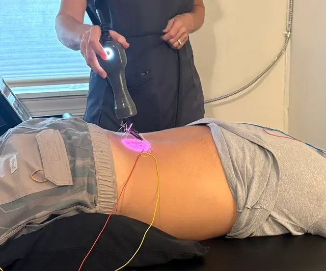 A practitioner in dark scrubs holds a laser wand over the lower back of a person lying on a treatment table