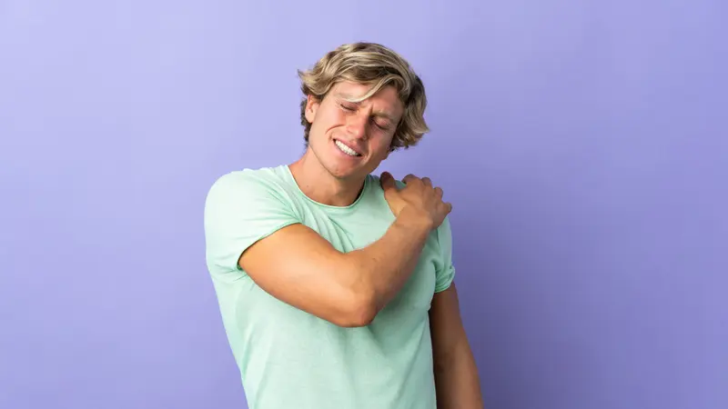 A young man in a green T-shirt holds his sore shoulder and winces
